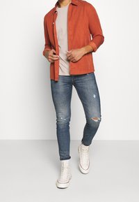 Rostfärgad skjorta med lång ärm, ljusgrå t-shirt, slitna blå jeans och vita högstövel sneakers, med en klassisk passform.