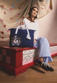 Mėlynos drobės rankinė su siuvinėtomis grafikos detalėmis, dvi šviesiai mėlynos rankenos ir raktų pakabukas su riedlenčių bateliu. Modelis pozuoja ant spalvoto pjedestalo.