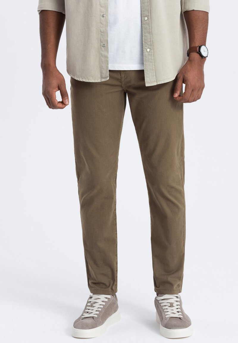 Chino kaki en tissu léger, coupe droite, avec des poches avant. Associés à des baskets en daim grises, lacets blancs, et une chemise à boutons.