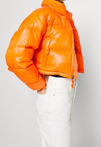 Skórzana kurtka puchowa w kolorze błyszczącej pomarańczy, o krótkim kroju, z wysokim kołnierzem, zamek błyskawiczny i dwoma bocznymi kieszeniami. Noszona na białych dżinsach.