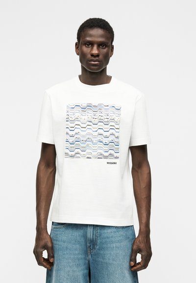 Άνδρας που φοράει λευκό T-shirt με τετράγωνο στο οποίο υπάρχει μπλε ζιγκ-ζαγκ μοτίβο και κείμενο «MISSONI», συνδυασμένο με ανοιχτό μπλε τζιν σορτς.