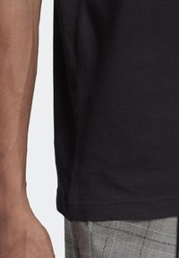 Černé bavlněné tričko s klasickým kulatým výstřihem, krátkými rukávy a volným střihem, v kombinaci se šedými kostkovanými kalhotami.