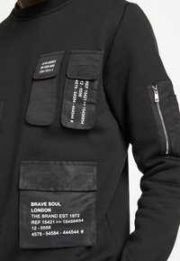 Černá mikina s více texturovanými kapsami a potištěnými detaily. Má zipovou kapsu na rukávu a značkování na spodní kapse.