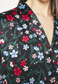 Blusa nera floreale con colletto a revers, dettagli con bottoni e un motivo di fiori rossi, blu e bianchi con foglie verdi. Tessuto morbido.