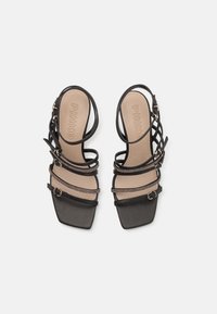 Sandale cu toc negru, cu vârf pătrat, având mai multe curele înguste și un accent metalic pe instep, pe o talpă de culoare bej.