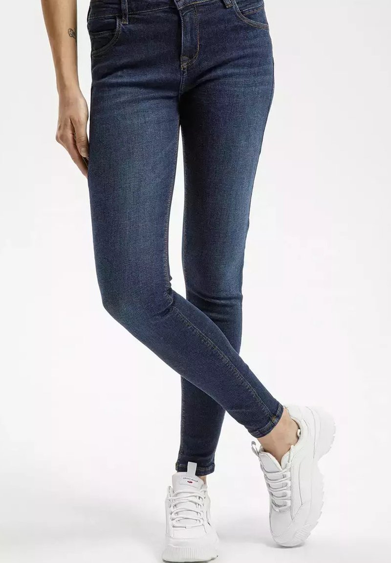 Chude dżinsy w ciemnoniebieskim denimu z delikatnymi detalami w szyciu, w połączeniu z białymi trampkami z teksturowaną podeszwą i sznurówkami.