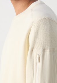 Krémový pulovr s texturovaným pletením a kontrastem hladké látky. Na levém rukávu je kapsa se zipem. Švové detaily přidávají rozměr.