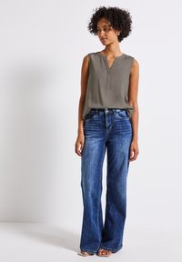 Ärmellose olivgrüne Bluse mit einem Einschlaghals, kombiniert mit hellblauen, weit geschnittenen Jeans. Das Outfit zeichnet sich durch einen lässigen, entspannten Stil und minimalen Hardware-Einsatz aus.