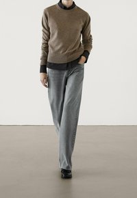 Maglione a maglia marrone sopra una camicia nera con bottoni, abbinato a jeans dritti grigi chiari e scarpe nere. Design semplice e lineare.