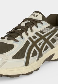 Καφέ και μπεζ παπούτσι τρεξίματος Asics με πλέγμα υφάσματος, αντανακλαστικές ασημί λεπτομέρειες, παχιά σόλα και δεμένα κορδόνια.