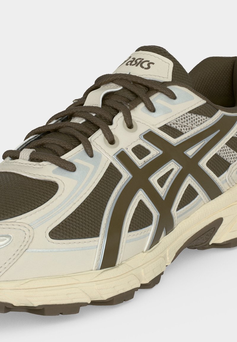 Zapatilla de correr Asics marrón y beige con tejido de malla, detalles plateados reflectantes, suela gruesa y cordones atados.