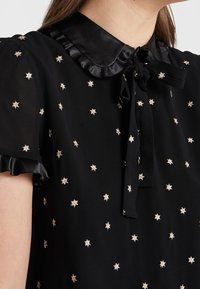Blusa nera con colletto arricciato in satin, motivi a stella in ricamo dorato e dettagli al collo annodati. Maniche corte con rifiniture arricciate.