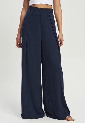 Marineblaue Hose mit weitem Bein und Falten, hoher Taille, getragen mit weißem kurz geschnittenem Top und beigen Riemchensandalen, gezeigt von der Taille bis zu den Füßen.