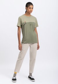 T-shirt in cotone verde oliva con design grafico, abbinata a pantaloni color crema. Sneakers nere con lacci bianchi, calzini grigi e dettagli gialli.