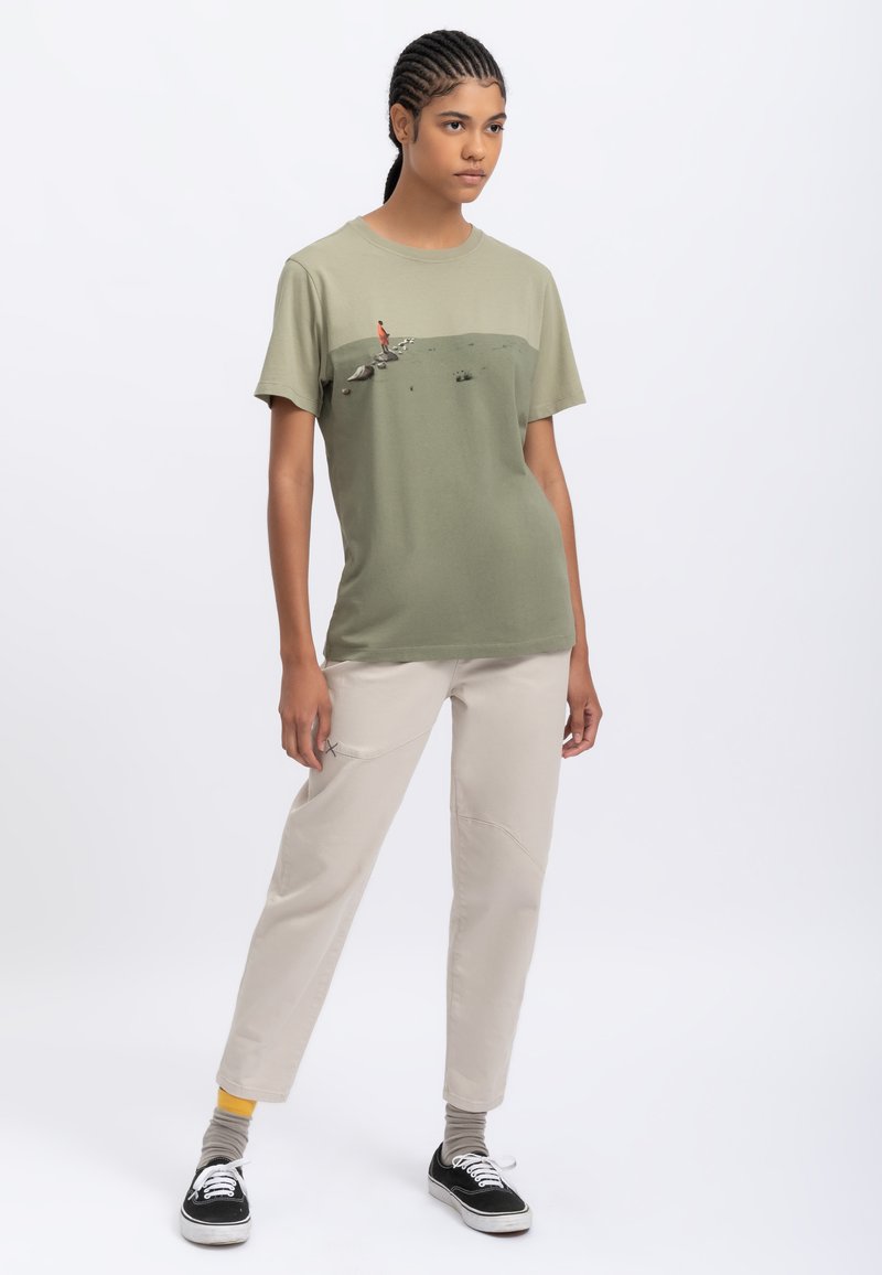 T-shirt in cotone verde oliva con design grafico, abbinata a pantaloni color crema. Sneakers nere con lacci bianchi, calzini grigi e dettagli gialli.