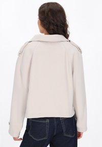Vaalean beige takki, jossa on kaulus, hartiatoppaukset ja pitkät hihat. Ominaisuuksiin kuuluu lyhennetty malli ja sileä, teksturoitu kangas.