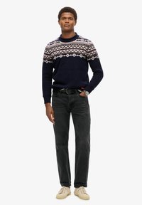 Marineblauw trui met geometrisch wit en rood patroon, gedragen met zwarte jeans en lichtbeige sneakers. Model staat rechtop, met de handen aan de zijkant.