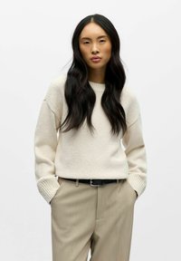 Kremfarget, teksturert strikkegenser med rund hals og ribbestrikkede mansjetter, kombinert med lysebeige bukser med folder og et svart belte.