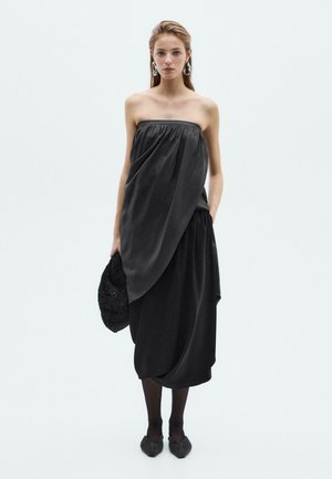 Abito nero senza spalline con tessuto drappeggiato, design asimmetrico e tasche. Indossato con una pochette nera testurizzata e ballerine con punta.