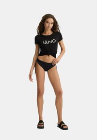 T-shirt nera a maniche corte con nodo davanti, caratterizzata dal logo "LIU JO" in un font decorativo. Abbinala a bikini bottom neri e sandali.