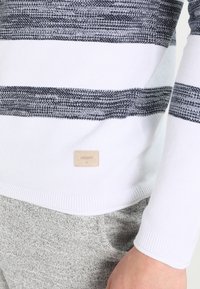 Pull en maille avec larges rayures horizontales noires et blanches, ourlet et manches côtelés, petite étiquette beige, porté avec un pantalon gris clair.