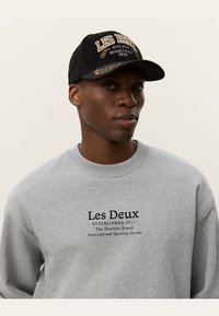 Felpa grigia con testo nero, con logo "Les Deux", abbinata a un cappello in corduroy nero, con dettagli in oro e ricami.