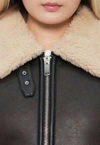 Primer plano de una persona que lleva una chaqueta de cuero negra con un cuello de borreguillo beige y una cremallera plateada parcialmente cerrada.