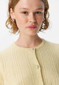 Gul stickad cardigan med rund halsringning, med kabelmönster och en pärlformad knapp i mitten. Mjuk textur, lätt material.