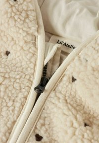 Veste en polaire couleur crème avec un extérieur texturé et duveteux, fermeture éclair et doublure beige. De petits points marron ajoutent des détails. Étiquette de marque visible.