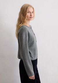 Femme blonde portant un gilet gris à boutons et un pantalon noir, se tenant de côté, regardant l'appareil photo devant un mur blanc uni.