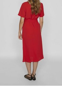 Robe midi rouge à manches courtes, taille cintrée et jupe fluide. Stylée avec des ballerines noires en mesh. Tissu lisse avec un drapé subtil.