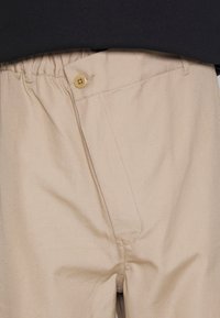 Pantalon beige avec taille élastique et fermeture à rabat boutonné, porté avec un haut noir.