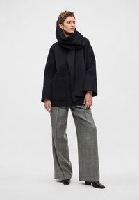 Černý vlněný vršek s vysokým obepínacím límcem a přední kapsou. V kombinaci se širokými šedými kostkovanými kalhotami a černými botami.