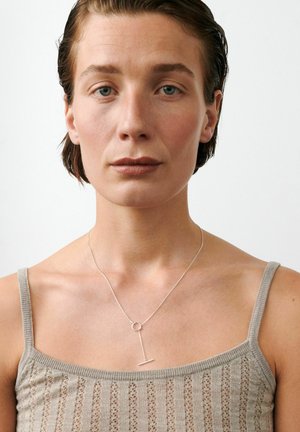 Žena s kratkom smeđom kosom koja nosi bež pleteni top na bretele i delikatnu srebrnu ogrlicu s okruglom kopčom i privjeskom.