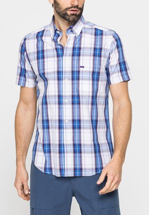Uomo che indossa una camicia a maniche corte a quadri blu, bianca e viola, abbinata a pantaloni blu scuro, in piedi davanti a uno sfondo semplice.