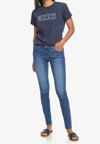 T-paita tummansinisestä puuvillasta, jossa on "ROXY"-logo, yhdistettynä slim-fit vaalean sinisiin farkkuihin ja mustiin liukuviin sandaaleihin.