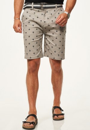 Mann trägt beige Shorts mit schwarzem Palmenmuster, gestreiften Gürtel, schwarze Sandalen, schwarzes Hemd, Armbanduhr und Armband, steht mit den Händen in der Nähe der Taschen.