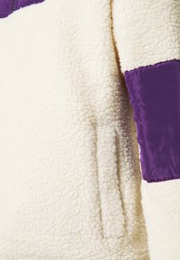 Kurtka z polaru z fakturowanym kremowym korpusem i fioletowymi nylonowymi akcentami na ramieniu i rękawie, z detalem bocznej kieszeni.
