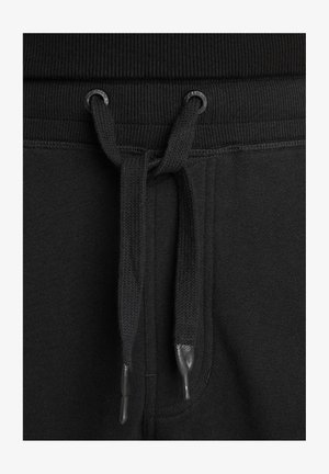 Acercamiento del cinturón de pantalones de chándal negros con un cordón atado a través de ojales metálicos negros y terminales de plástico en los extremos del cordón.