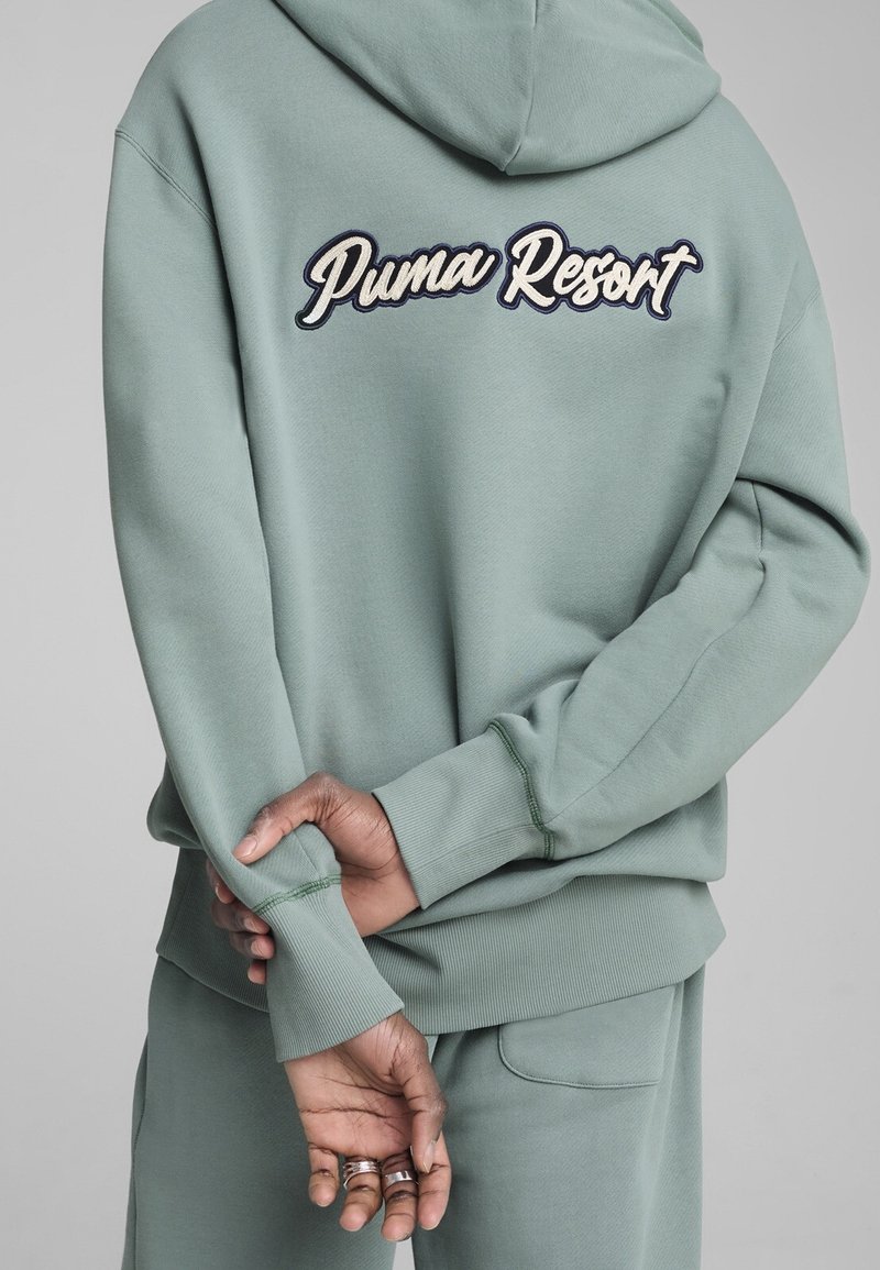 Persoon draagt een lichtgroene hoodie en joggingbroek met 'Puma Resort' geborduurd op de achterkant, handen achterrug in elkaar gevouwen.