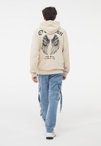 Felpa beige con grafica di ali nera e testo sul retro. Abbinata a pantaloni cargo azzurri chiari con molteplici tasche e lacci all'orlo.