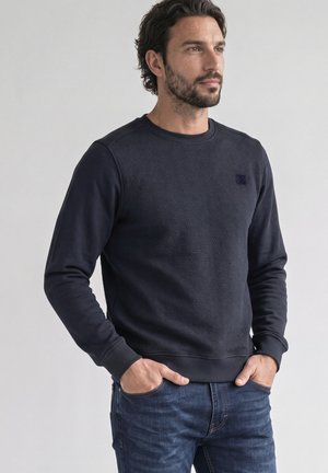 Mann mit dunklen Haaren und Bart trägt einen marineblauen, strukturierten Pullover und blaue Jeans, steht mit den Händen in den Taschen vor einem schlichten Hintergrund.
