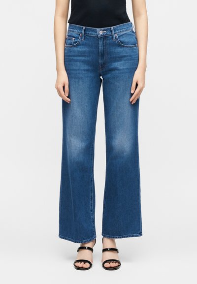 Γυναίκα που φοράει μπλε τζιν με ψηλή μέση και φαρδιά πόδια, σε συνδυασμό με μαύρα σανδάλια με λουράκια, στέκεται μπροστά σε μια απλή λευκή επιφάνεια.