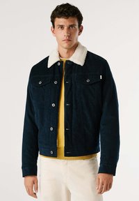 Giacca in velluto a coste blu navy con colletto in pelliccia sintetica bianca, dotata di due tasche frontali, bottoni metallici e indossata sopra un maglione giallo.