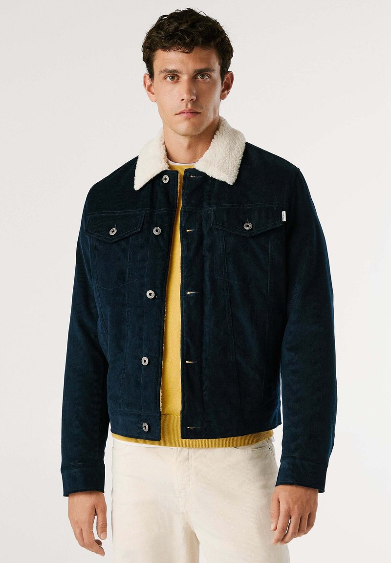 Giacca in velluto a coste blu navy con colletto in pelliccia sintetica bianca, dotata di due tasche frontali, bottoni metallici e indossata sopra un maglione giallo.