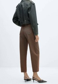 Žena v krátké černé kožené bundě, s vysokým pasem hnědých kalhotách a černých lodičkách s špičatou špičkou, stojící na bílém pozadí.