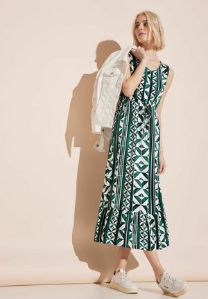 Green patterned maxi dress with geometric designs, sleeveless bodice, cinched waist, and ruffled hem, paired with a white denim jacket and sneakers.