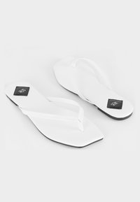 Witte flip-flops met een glanzende afwerking, met een platte zool en een geometrische, vierkante vorm. Zwart logo op een rechthoekig accent.