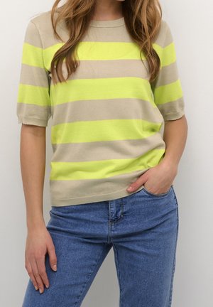Kvinde iført en beige og neon gul kortærmet trøje med vandrette striber og blå jeans, hånd i lommen, foran en ensfarvet lys baggrund.