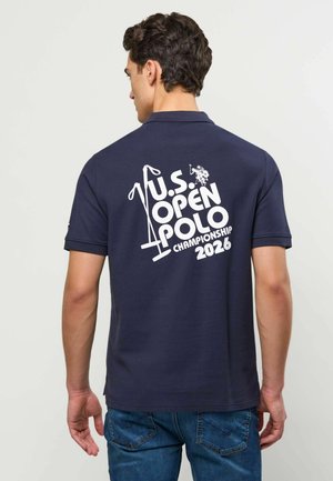 Férfi sötétkék galléros pólót visel, melyen fehér "U.S. Open Polo Championship 2026" logó és pólojátékos grafika található a hátán, hátulról nézve.